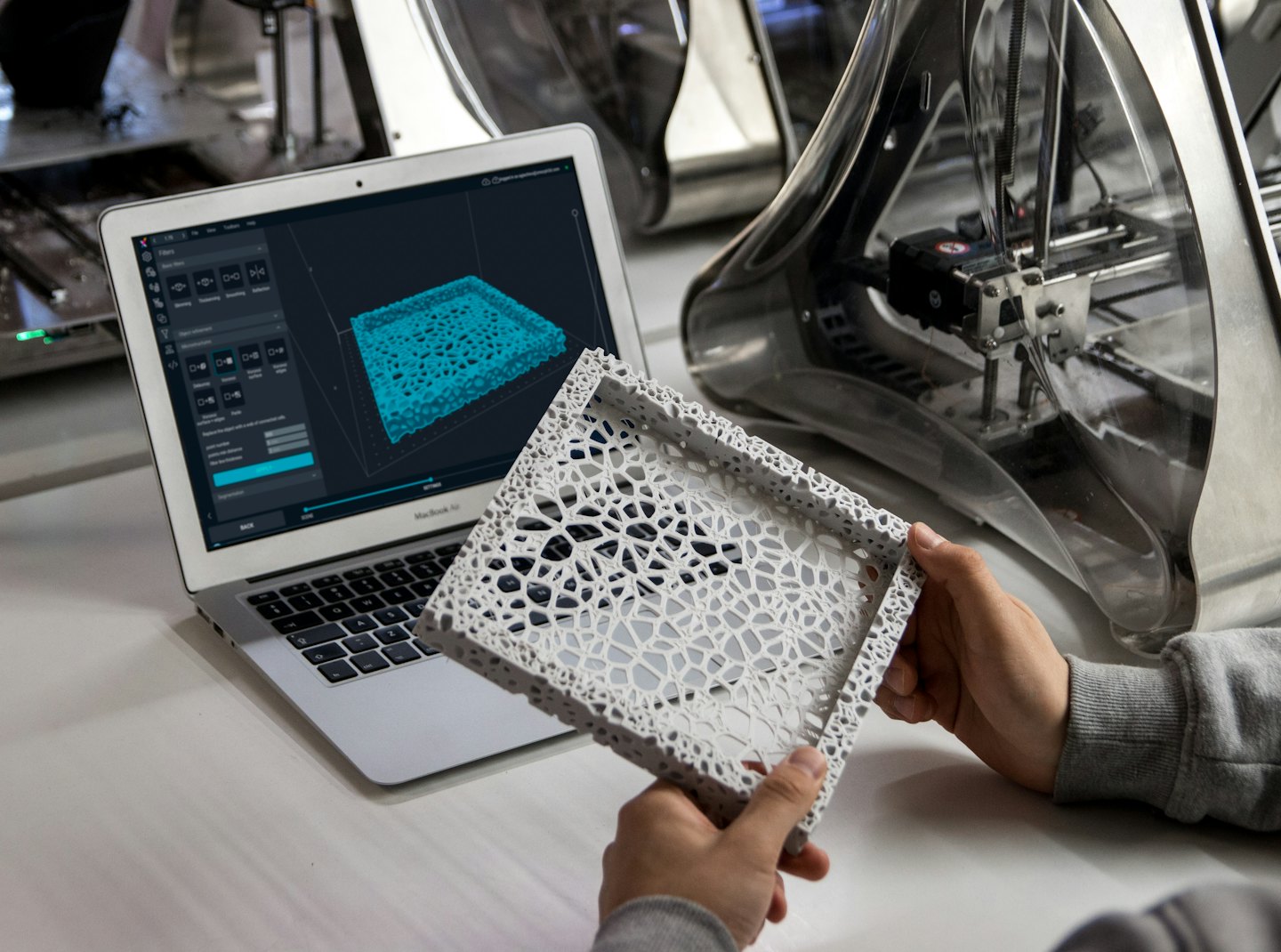Ruky držia 3D vytlačený objekt vedľa notebooku zobrazujúceho 3D model.